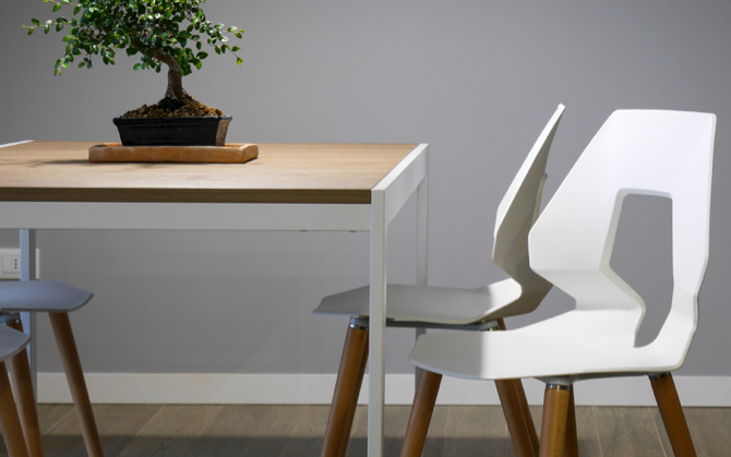 Two modern white chairs with angular cut out backs and wooden legs beside a wooden table topped with a miniature tree.